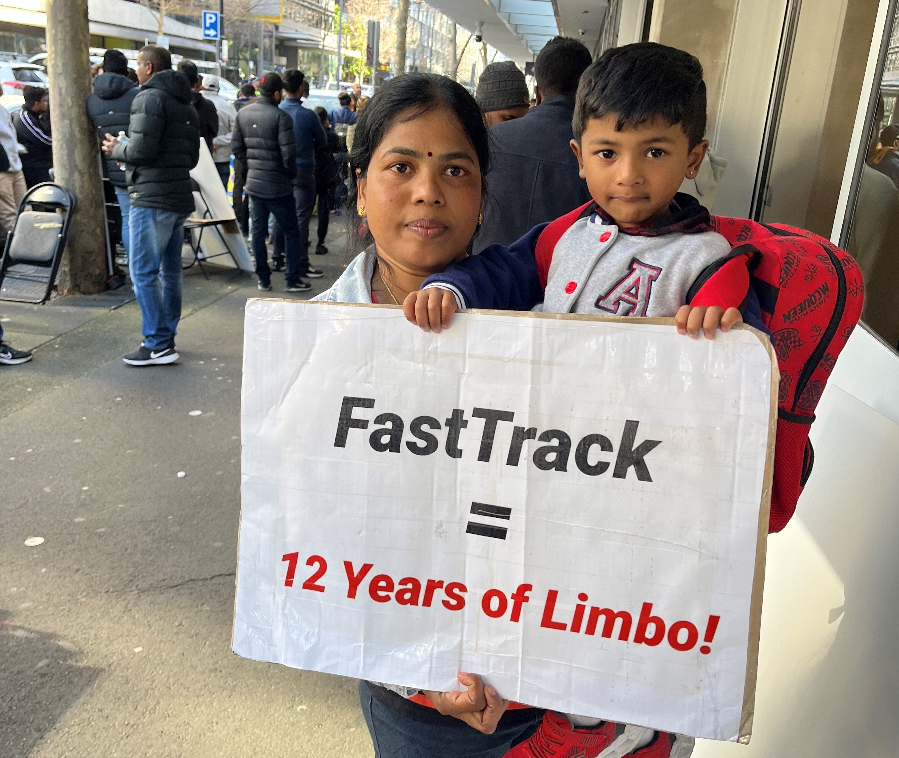 A woman stands holding her child and a placard that reads 'fasttrack = 12 years of limbo'