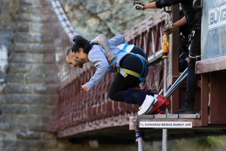 A woman and a man wearing safety kit hold each other as they tilt foward moments before jumping off a bungee-jump platform.