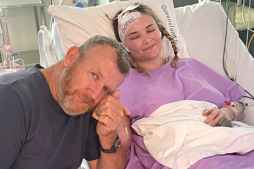 Dad and young adult daughter, she's laying in hospital bed with a bandage over head saying 'no bone'