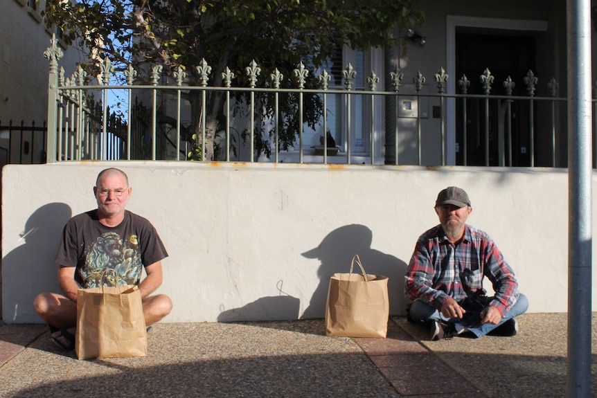 two men sitting on pavement - about two metres apart with paper bags in front of them