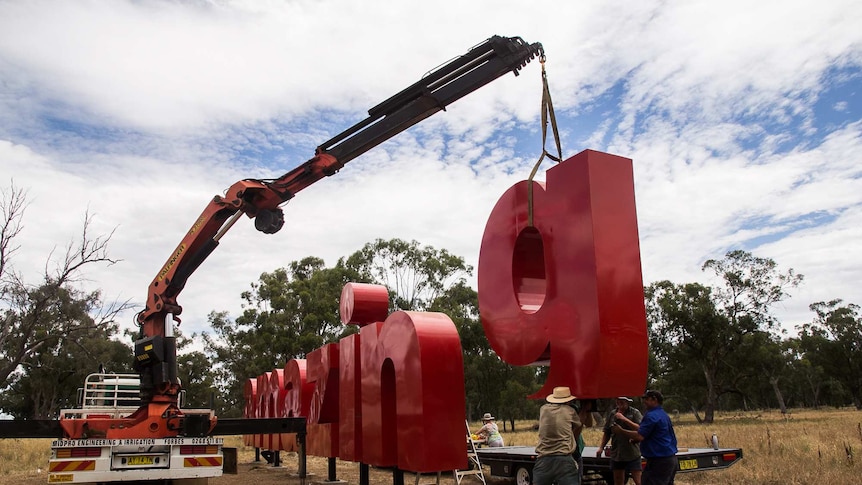 A crane lowering a red metal 'g' into place on pylons in a paddock.