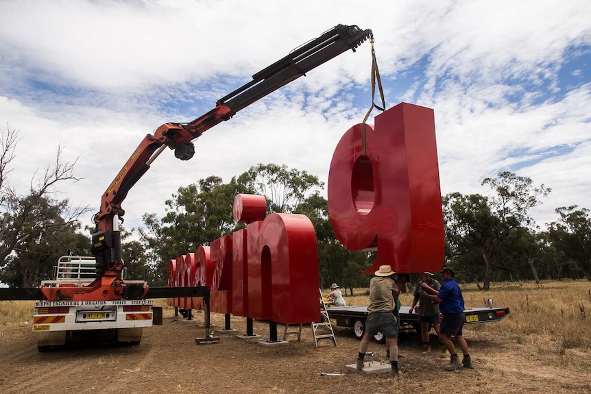 A crane lowering a red metal 'g' into place on pylons in a paddock.