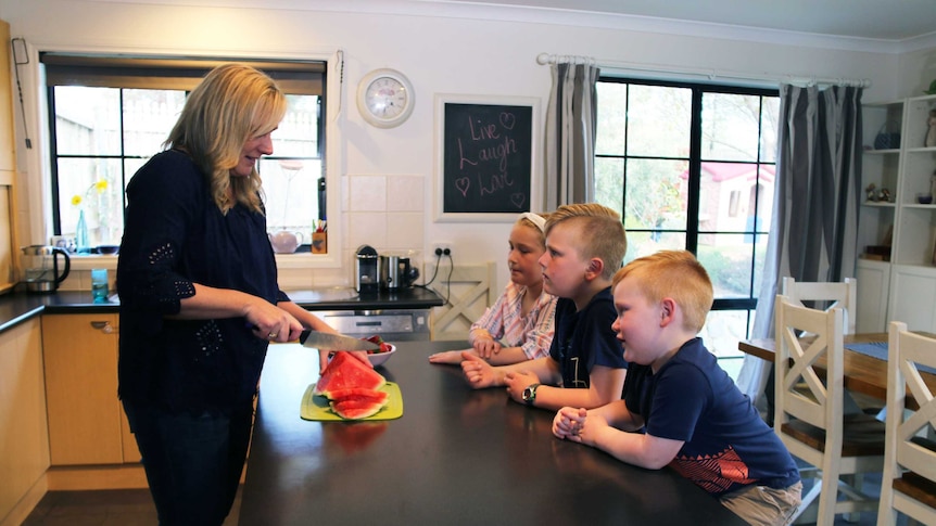 A woman cutting watermelon in a kitchen with three children looking at her from across a bench