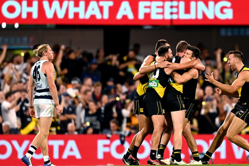 A group of victorious AFL players gather for a group hug after the final siren in grand final, as a dejected opponent looks on.