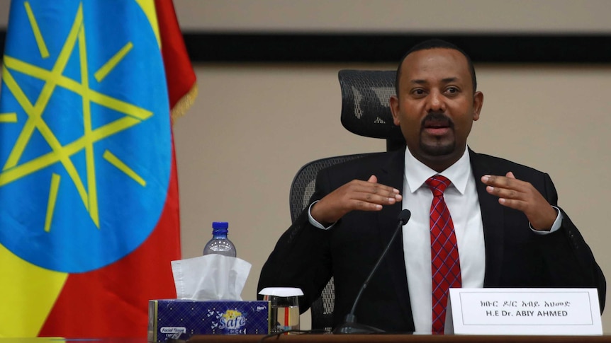 Man with dark hair, skin and goatee speaks at table with colourful flag in background.