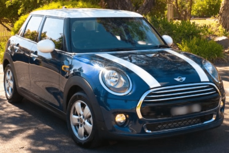 A dark blue coloured Mini Cooper car with two white stripes down the bonnet and a white roof parked under a tree on a sunny day.