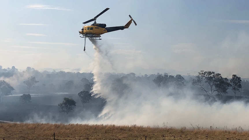 A blue and white helicopter drops water on a paddock dotted with trees as smoke billows.