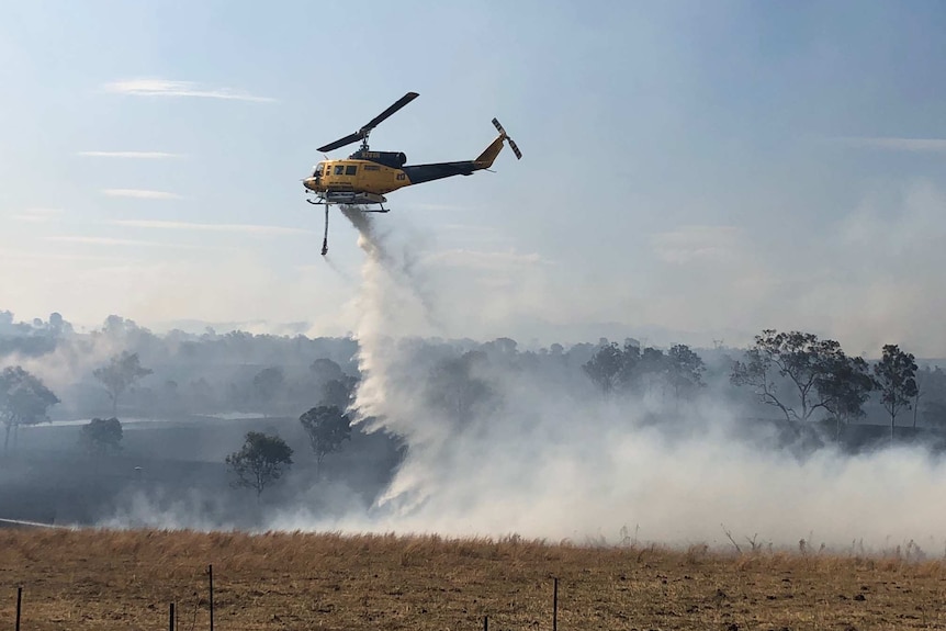 A blue and white helicopter drops water on a paddock dotted with trees as smoke billows.