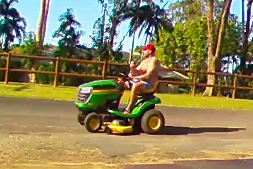 A man on a ride-on lawnmower with a beer in hand drive down a street in Cairns.