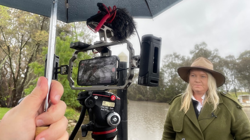 Woman wearing an akubra hat standing in front of flooded river looking at a camera under an umbrella.