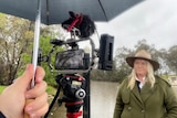 Woman wearing an akubra hat standing in front of flooded river looking at a camera under an umbrella.
