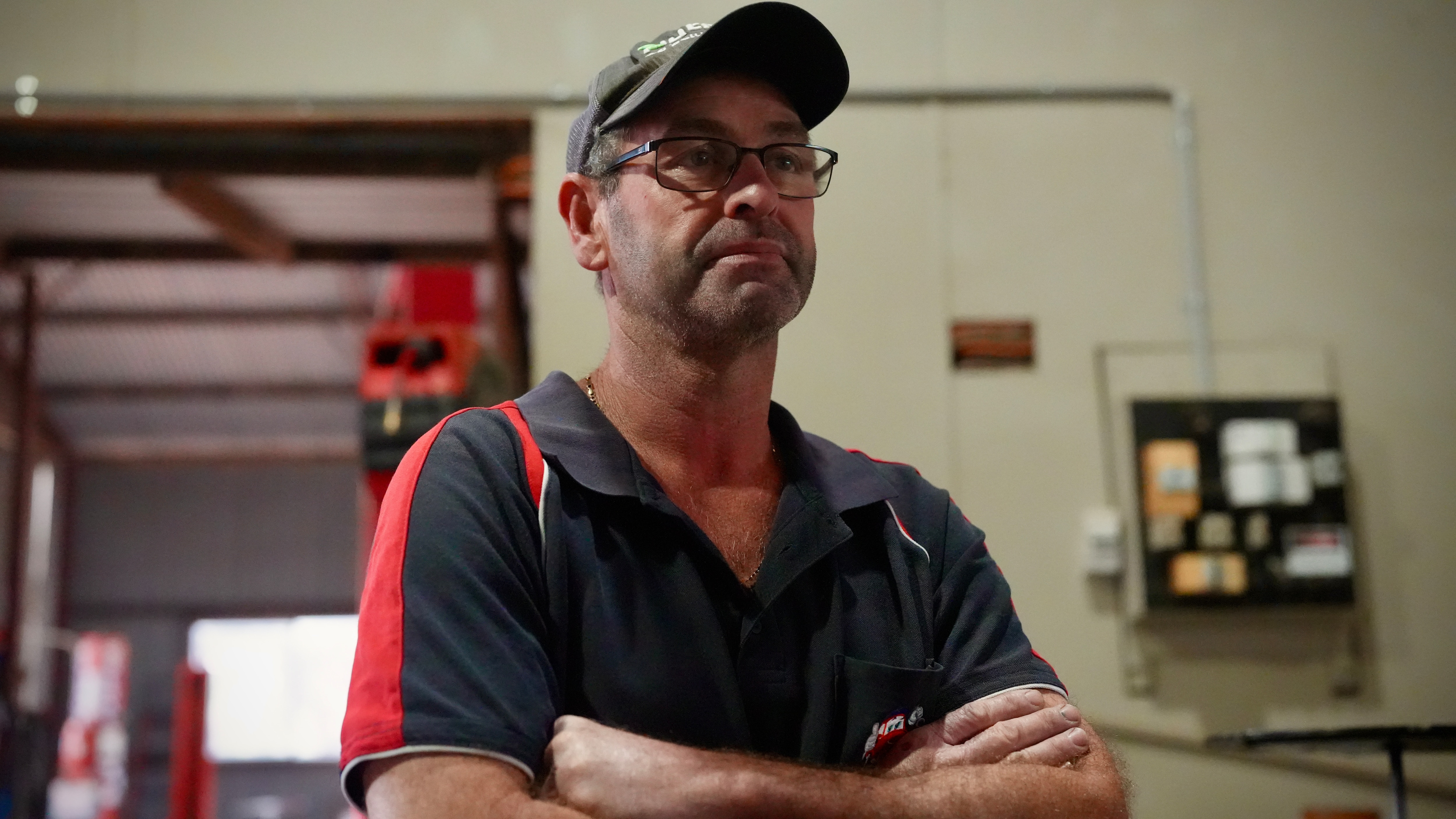 Un hombre de mediana edad que lleva una camisa de trabajo de mecánico negra y roja, gorra negra y gafas mira a la derecha del marco en el taller de mecánico.
