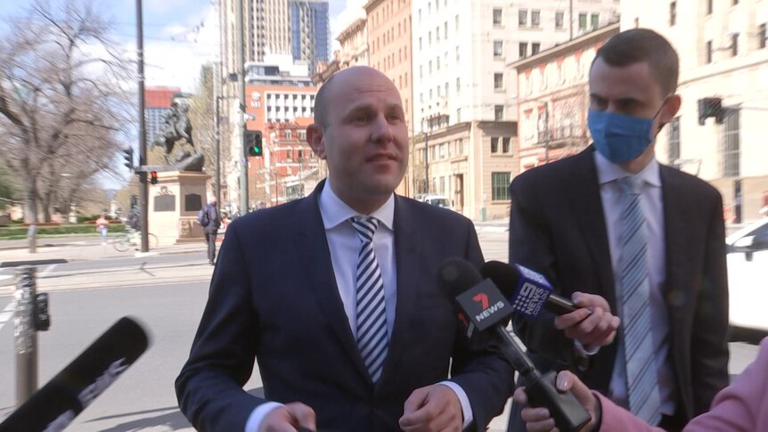 A bald man wearing a suit speaks to a journalist and other microphones outside
