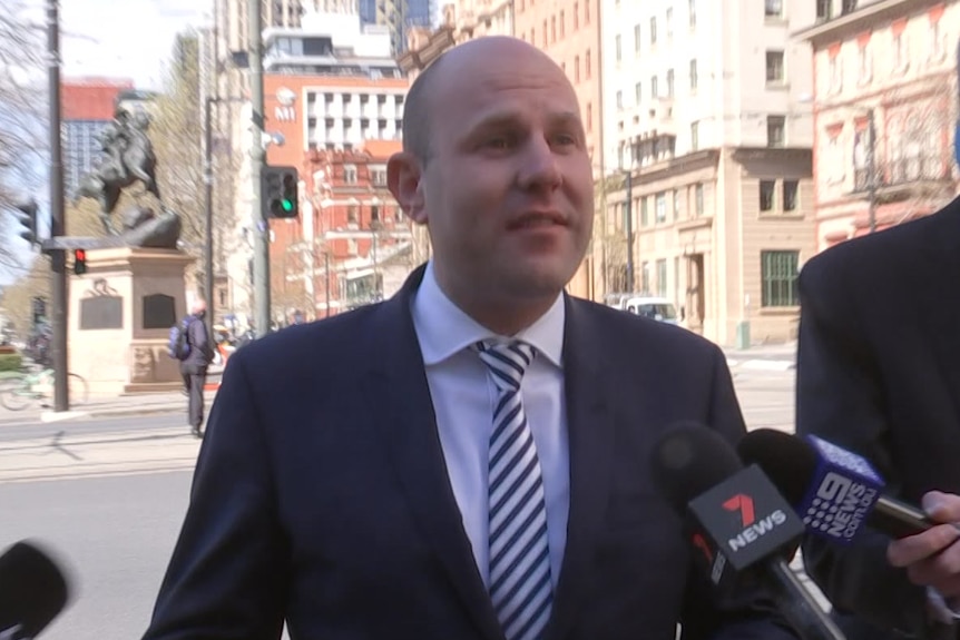 A bald man wearing a suit speaks to a journalist and other microphones outside