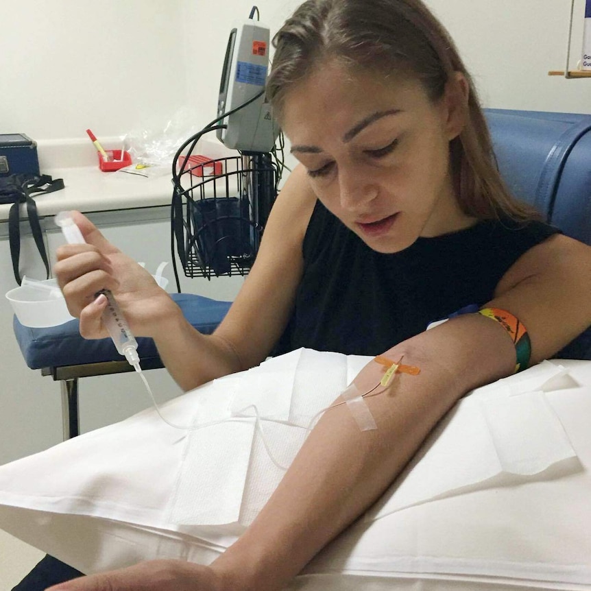 Chloe Christos sits in a chair with a needle in her arm and a syringe in her hand as she gets treatment for a blood disorder.