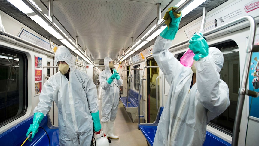 Three men wearing white overalls and face masks clean the inside of train with spray bottles.