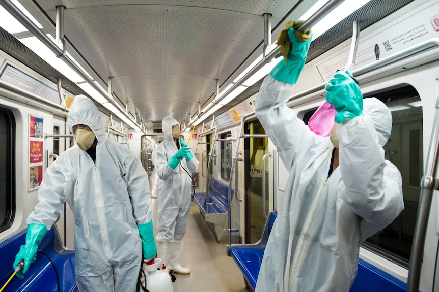 Three men wearing white overalls and face masks clean the inside of train with spray bottles.