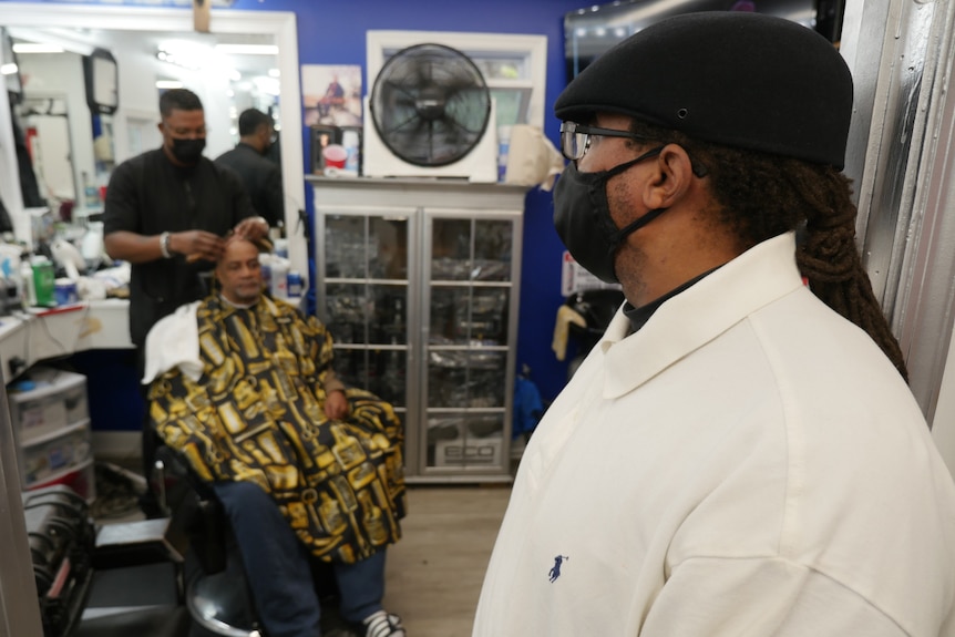 A man in a white shirt, black mask and black cap watching as a man cuts another man's hair in a barber shop