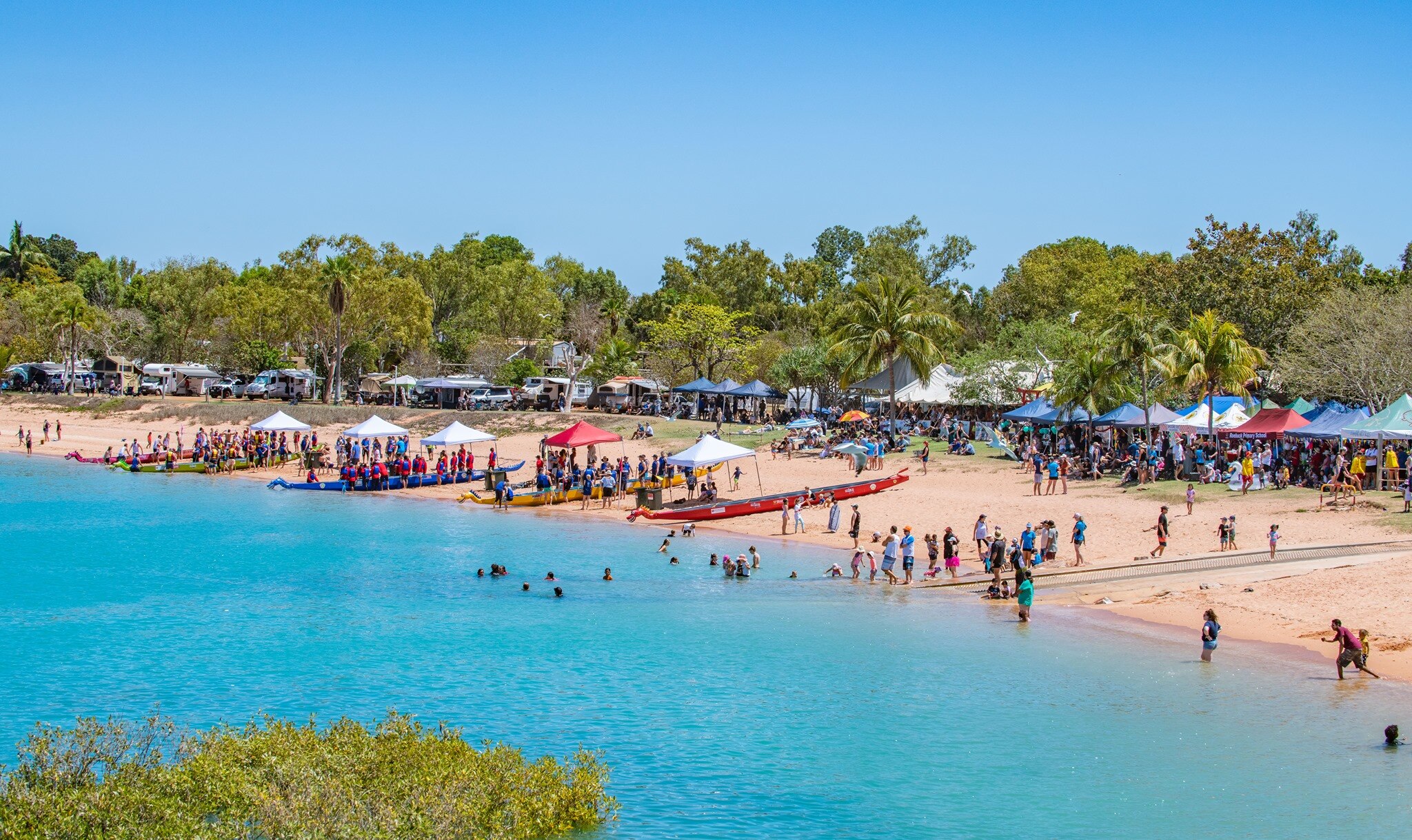 A wide shot of stalls, people and dragon boats lined up along turquoise waters of a bay.