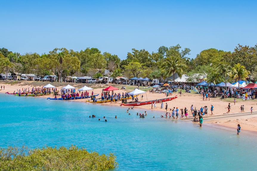 A wide shot of stalls, people and dragon boats lined up along turquoise waters of a bay.