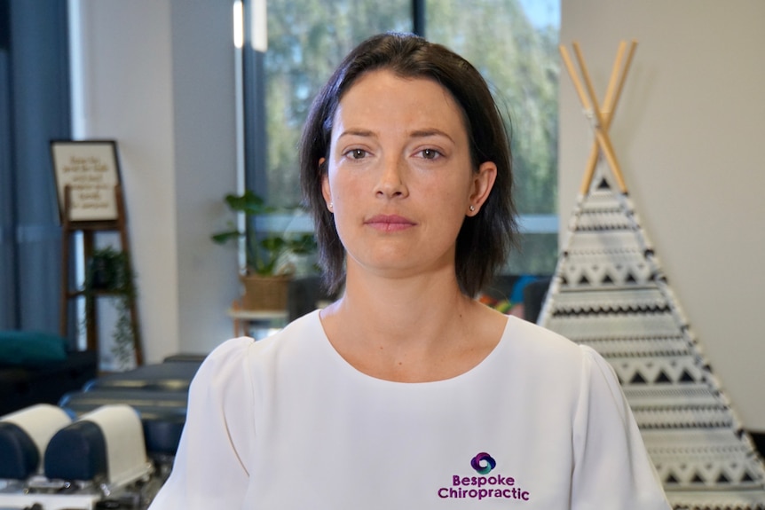 A dark-haired woman stands in a chiropractic clinic, with a treatment bed and an Indian tee-pee behind her