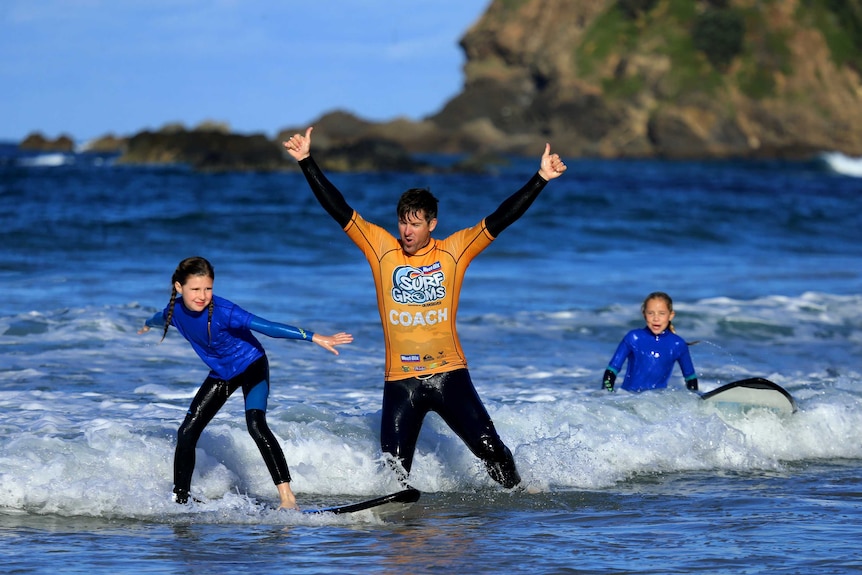 A young girl surfing a wave, with a surf coach near her with his arms in the air.