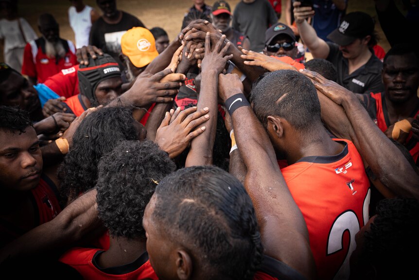 A group of players in orange jerseys standing in a circle, heads down, with their arms all reaching towards the centre