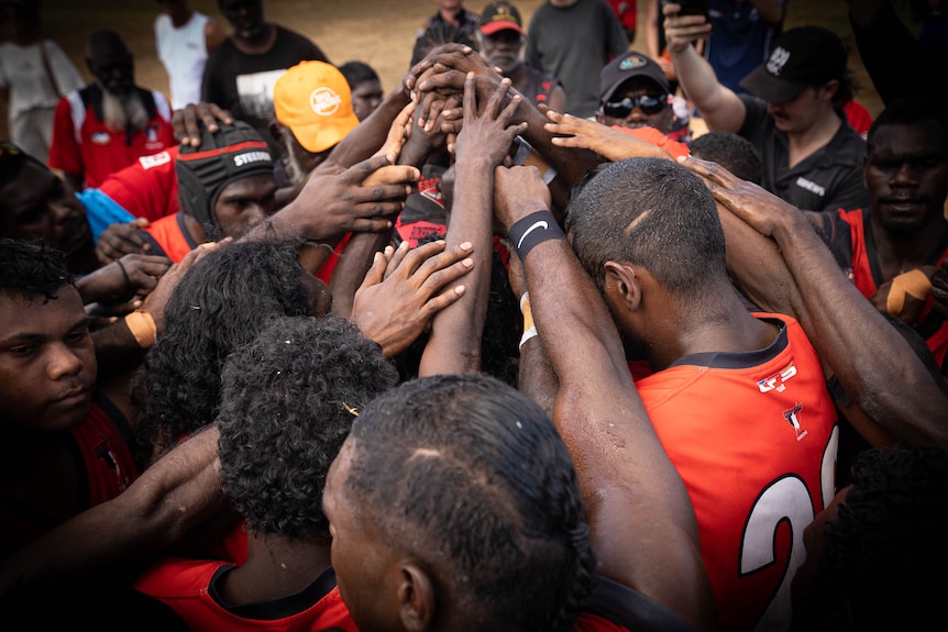 A group of players in orange jerseys standing in a circle, heads down, with their arms all reaching towards the centre