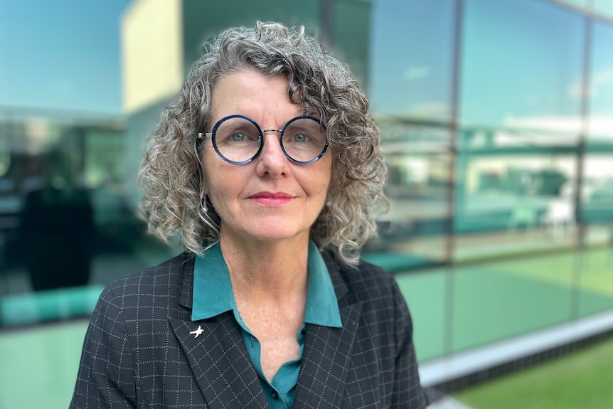 A portrait of Christina Pollard wearing thick-rimmed glasses, smiling in front of an office building.