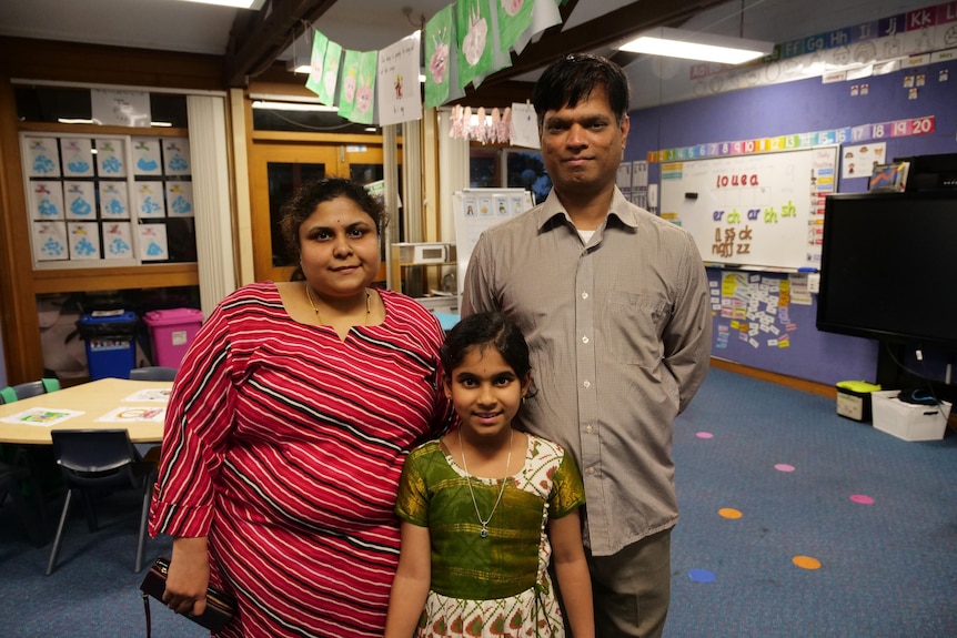 A family of three smiling standing in a primary school classroom