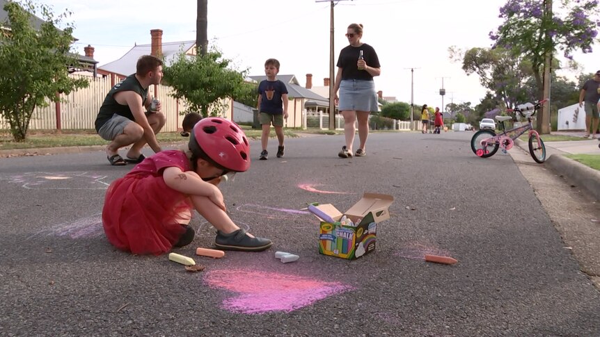 A child in a bike helmet chalk-drawing on the street with a man, a woman and a child in the background