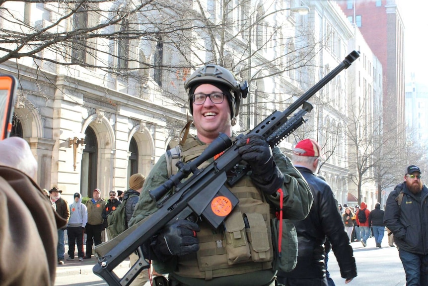 a man wearing glasses and camo and a helmet smiles as people take photos of him holding his rifle