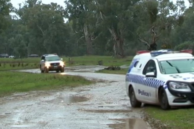 Two police cars driving on a muddy dirt track in a green, rural setting with trees in the background.