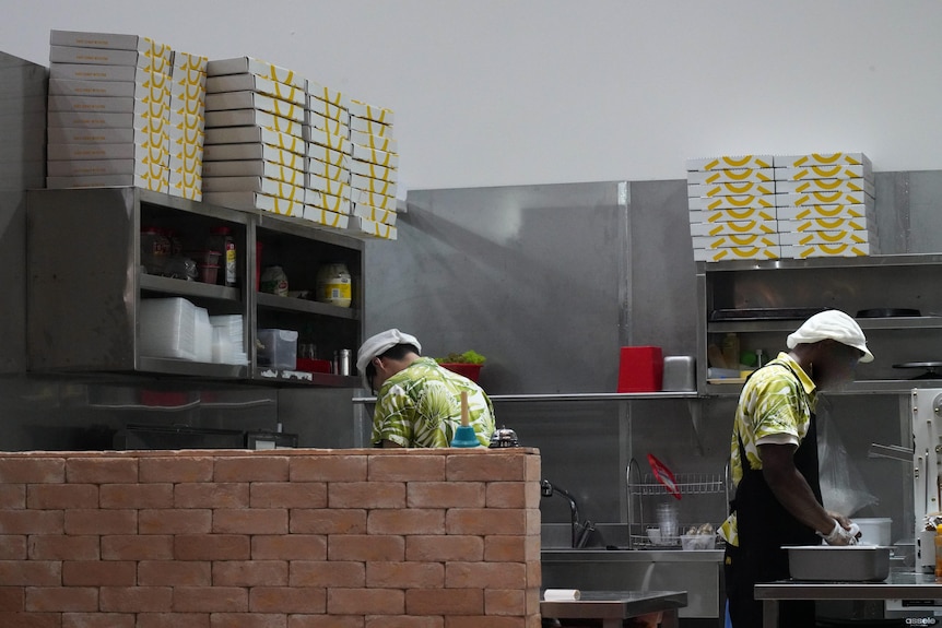 Two workers in a commercial kitchen with shelves stacked with pizza boxes. 