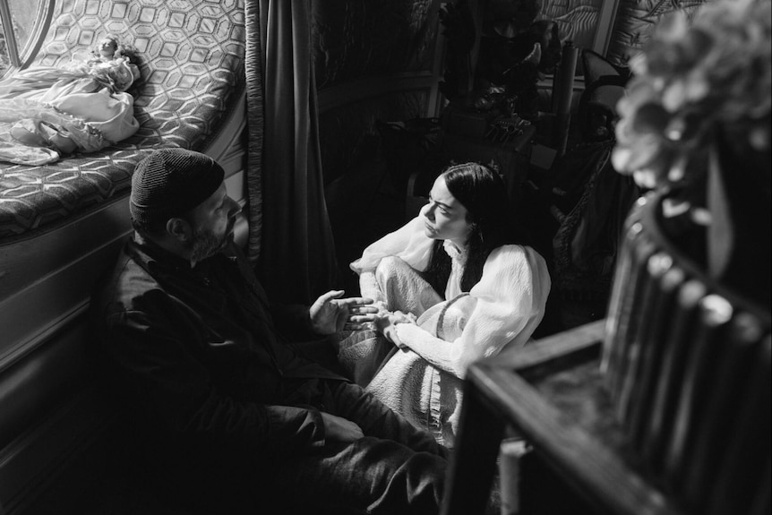 A black and white photo of Emma, a young woman with long black hair, sitting on the bedroom floor set with Yorgos.