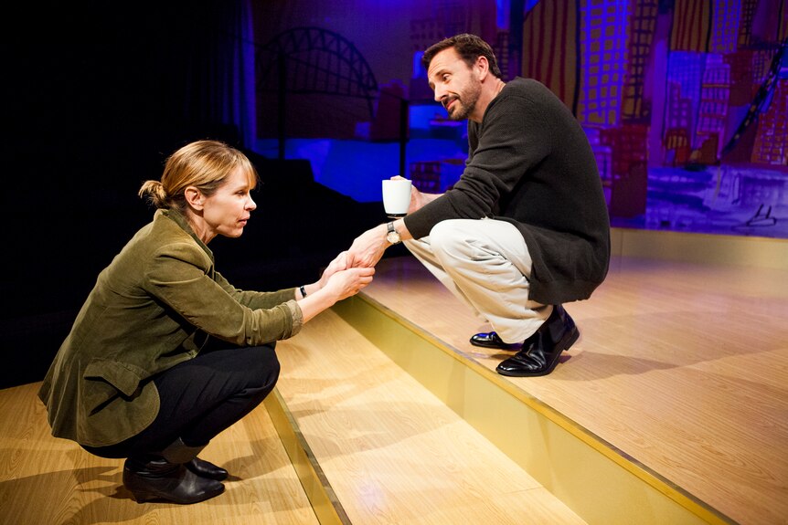 A middle-aged woman crouches at the foot of two steps on stage, holding a middle-aged man's hand with both hands.