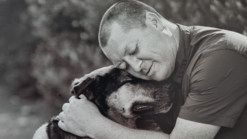 A middle-aged man with a shaved head cuddles his dog with his eyes closed.