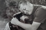 A middle-aged man with a shaved head cuddles his dog with his eyes closed.