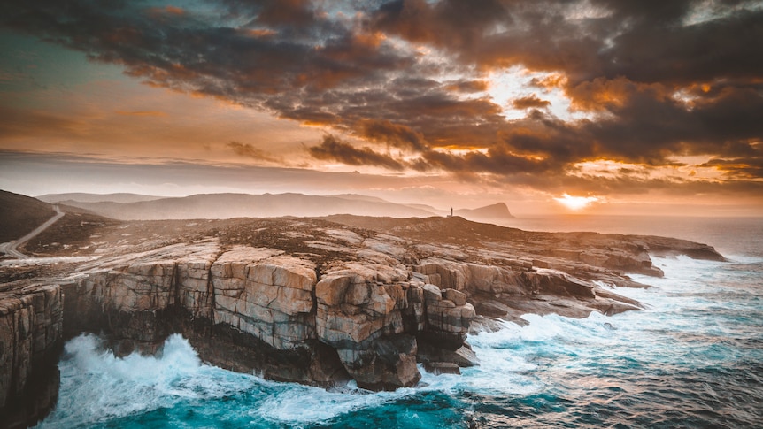 Rough seas smash against the rock walls of the albany coastline as the sun rises through clouds in the background