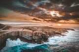 Rough seas smash against the rock walls of the albany coastline as the sun rises through clouds in the background