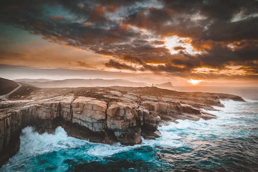 Rough seas smash against the rock walls of the albany coastline as the sun rises through clouds in the background