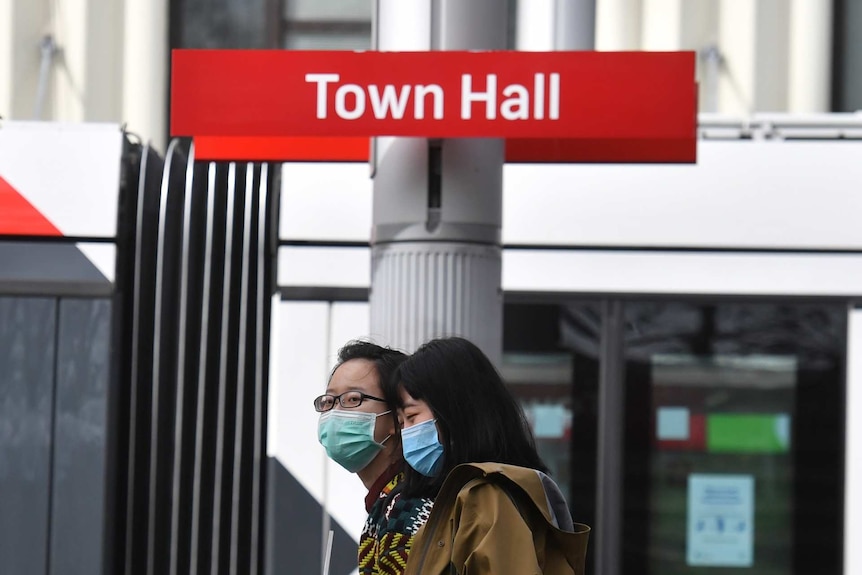 two women in face masks underneath a tram sign reading town hall