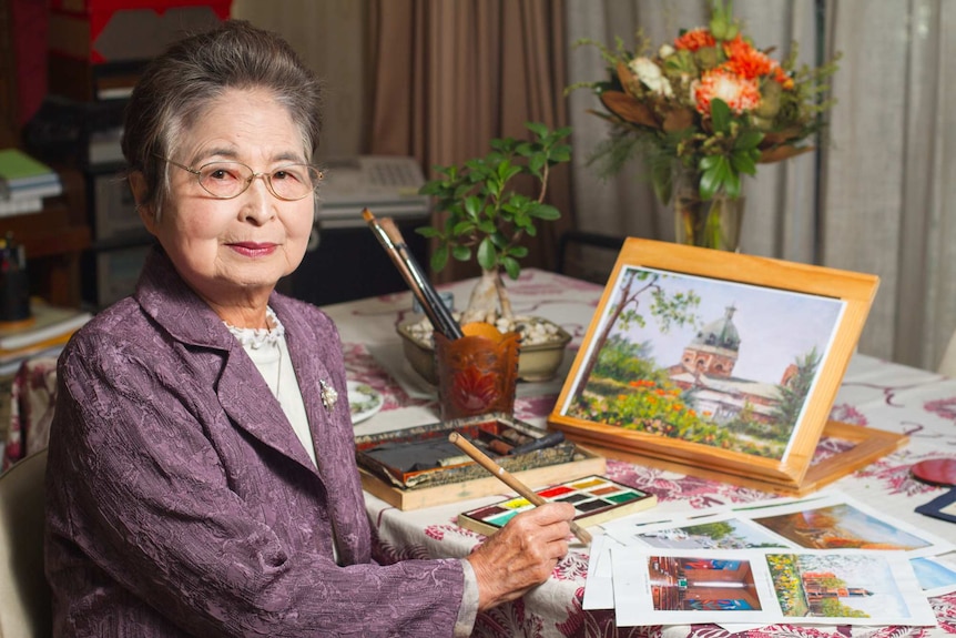 A woman sits at a table with brushes, paints and artworks around her
