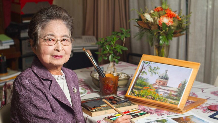 A woman sits at a table with brushes, paints and artworks around her