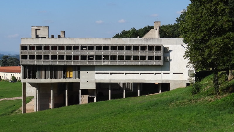 Looking across a sloping green hill, you view the brutalist shape of the La Tourette convent on a bright blue day.