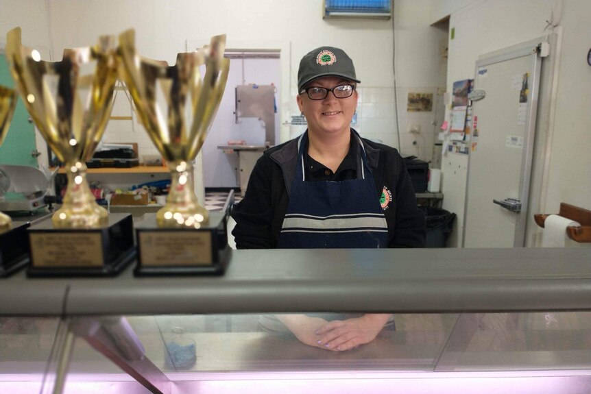A woman wears a dark shirt, a hat and glasses. She wears a striped apron, she stands in a shop front with gold trophies.