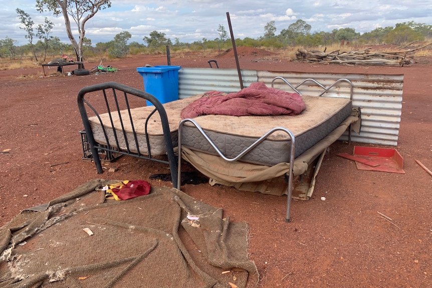 two mattresses on bed frames are shown next to a bin and sheets on red dirt outside