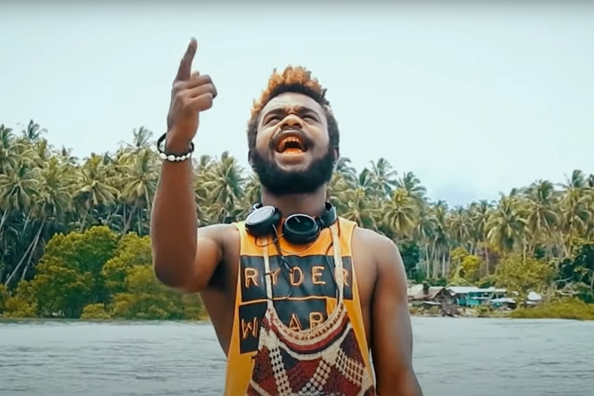 A Papua New Guinean man wearing singlet and headphones around neck points to the sky with island and water in background