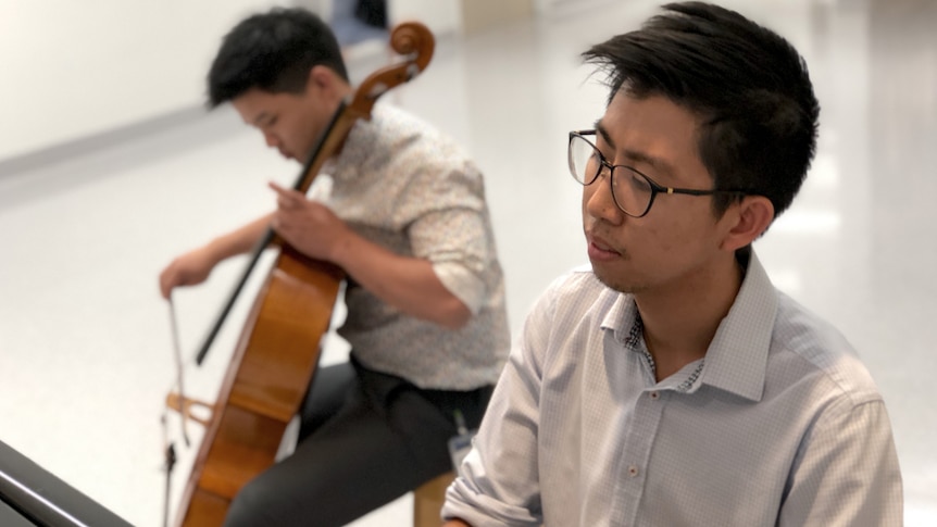 Two young men who are doctors and one plays the cello while the other plays piano in the hospital foyer.