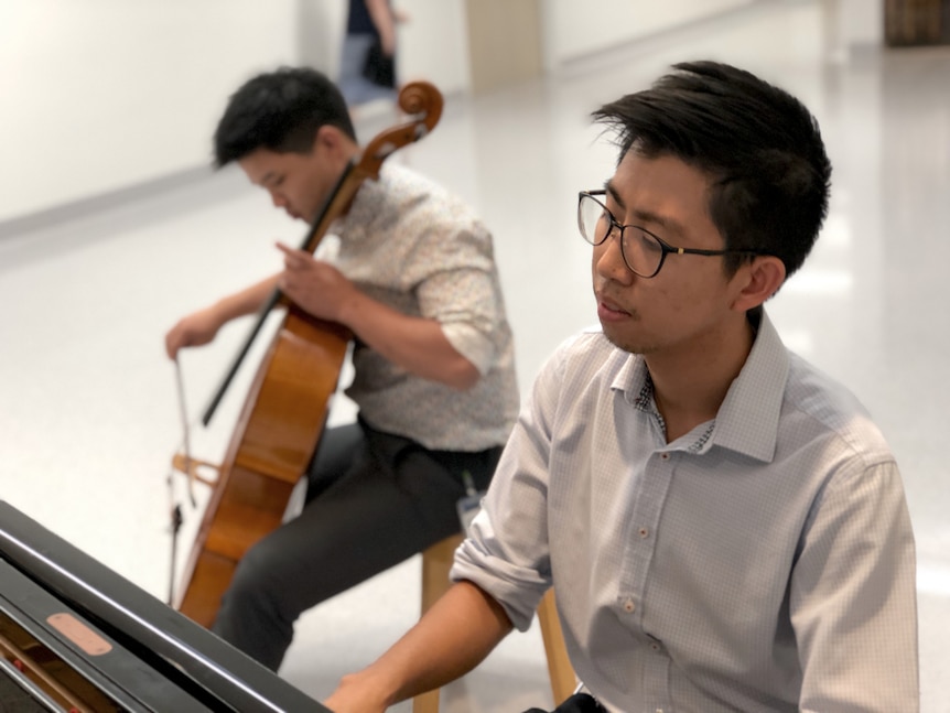 Two young men who are doctors and one plays the cello while the other plays piano in the hospital foyer.